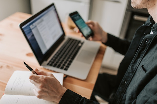 person holding a pen and their phone will looking at something on their laptop
